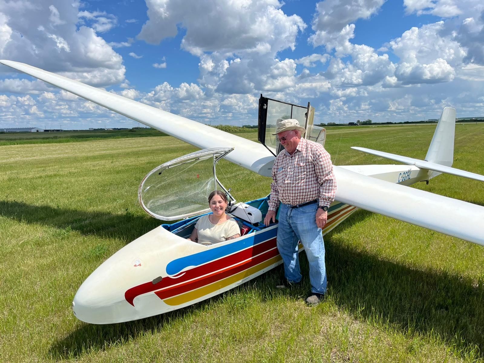 A young student sits in the front seat of the two-seat glider while a club member stands beside the cockpit